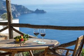 Una taula de fusta amb dues copes de vi i un plat de fruita a un balcó amb vista al mar.