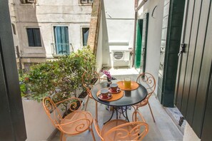 Un piccolo balcone esterno con tavolo e sedie, una pianta in vaso e vista su un edificio con una porta verde.