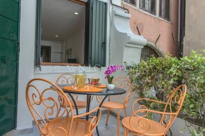 An outdoor seating area with orange metal chairs and a table, a potted plant, and a window with a view of the interior.