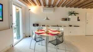 Un espace repas moderne avec une table ronde pour quatre personnes, des chaises transparentes, un luminaire suspendu et un plafond blanc avec des poutres apparentes.