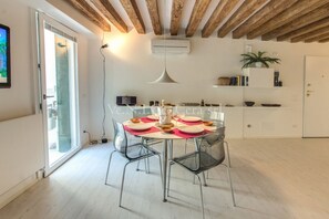 Un'elegante zona pranzo con un tavolo rotondo apparecchiato per quattro persone, sedie trasparenti, una lampada a sospensione e un soffitto bianco con travi a vista.