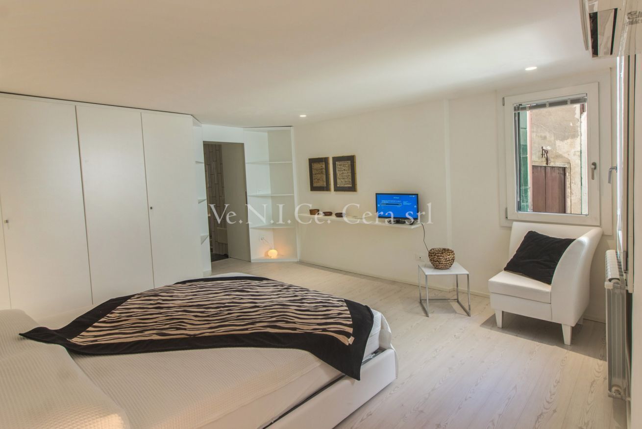 A modern bedroom with a bed, a white wardrobe, a small bedside table, and a chair.