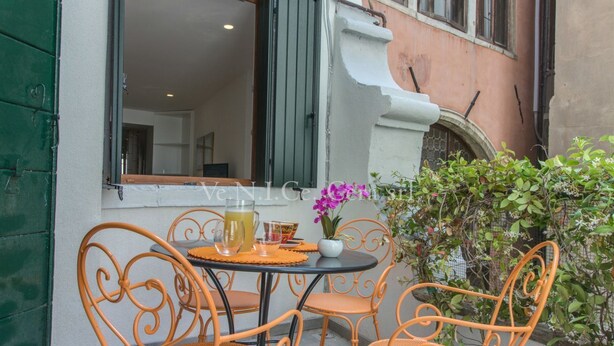 Un espace extérieur aménagé avec des chaises et une table en métal orange, un pot de fleurs et une fenêtre donnant sur l’intérieur.