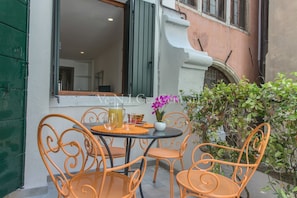 Un espace extérieur aménagé avec des chaises et une table en métal orange, un pot de fleurs et une fenêtre donnant sur l’intérieur.