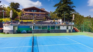 Een tennisbaan met een net in het midden, omgeven door een groen gebied en een gebouw met een balkon op de achtergrond.