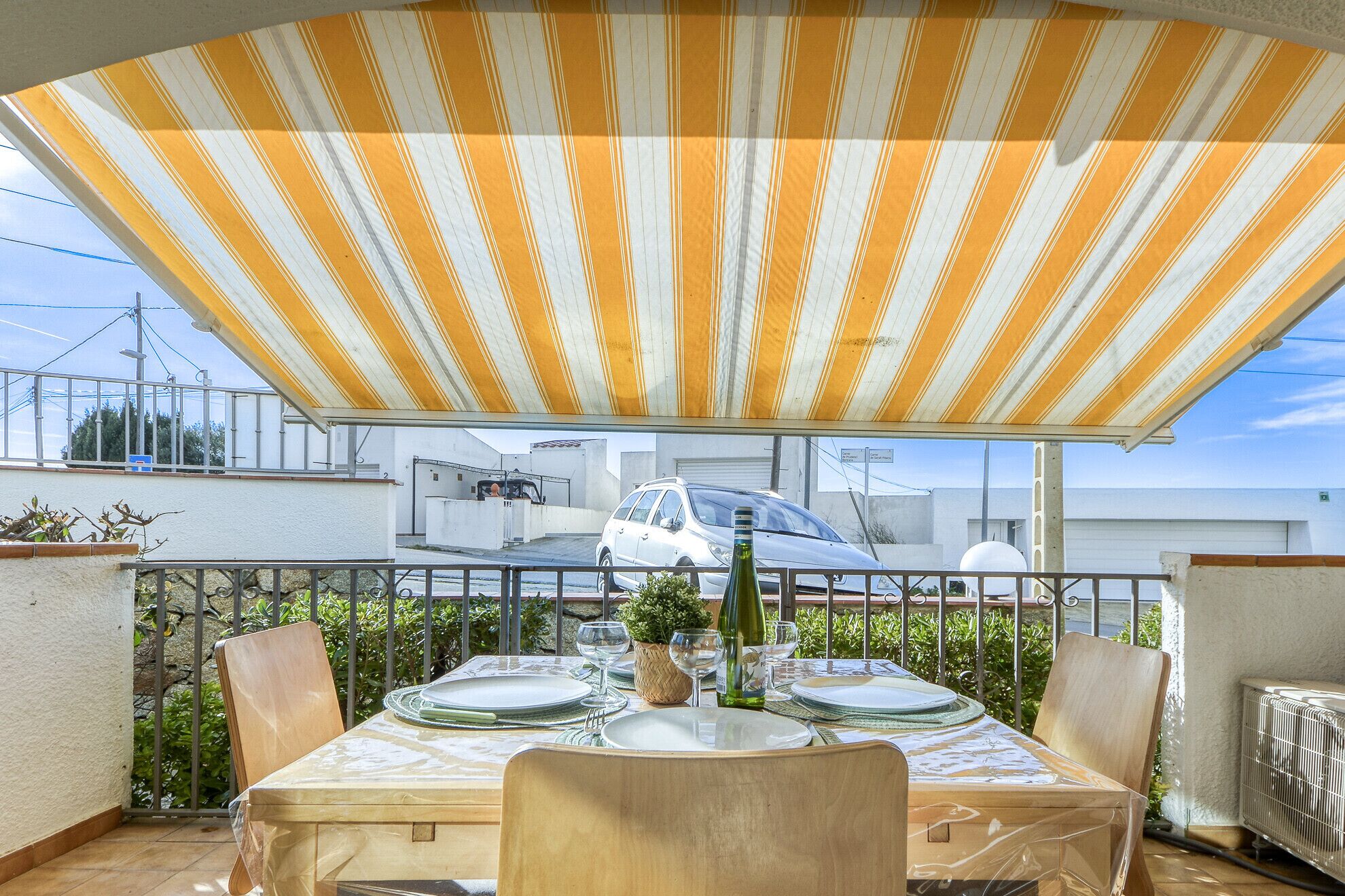 An outdoor patio with a striped awning, a table set for dining, and a view of parked cars and buildings in the background.