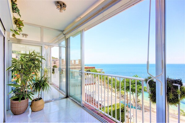 A balcony with a view of the sea, large potted plants, and a glass door leading to the interior.