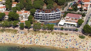 An aerial view of a beach with numerous umbrellas and people, a curved building with balconies, and a road nearby. - Beachfront two-bedroom apartment with sea views in Canyelles Petites (Roses)