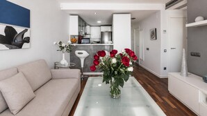 A modern living room with a sofa, a glass coffee table with a vase of flowers, a kitchen in the background, and a white wall with a blue artwork. - Beachfront two-bedroom apartment with sea views in Canyelles Petites (Roses)