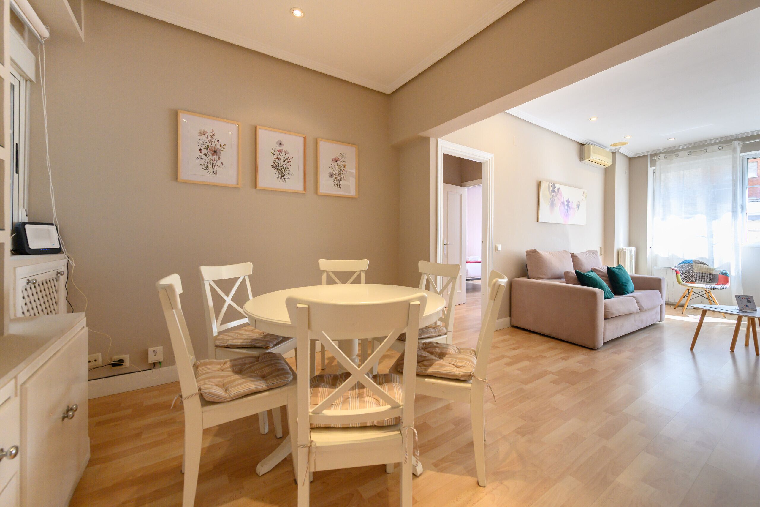 Un salon avec une table ronde, des chaises blanches, un canapé et une table basse. Des tableaux encadrés ornent le mur, et une fenêtre est agrémentée de rideaux.