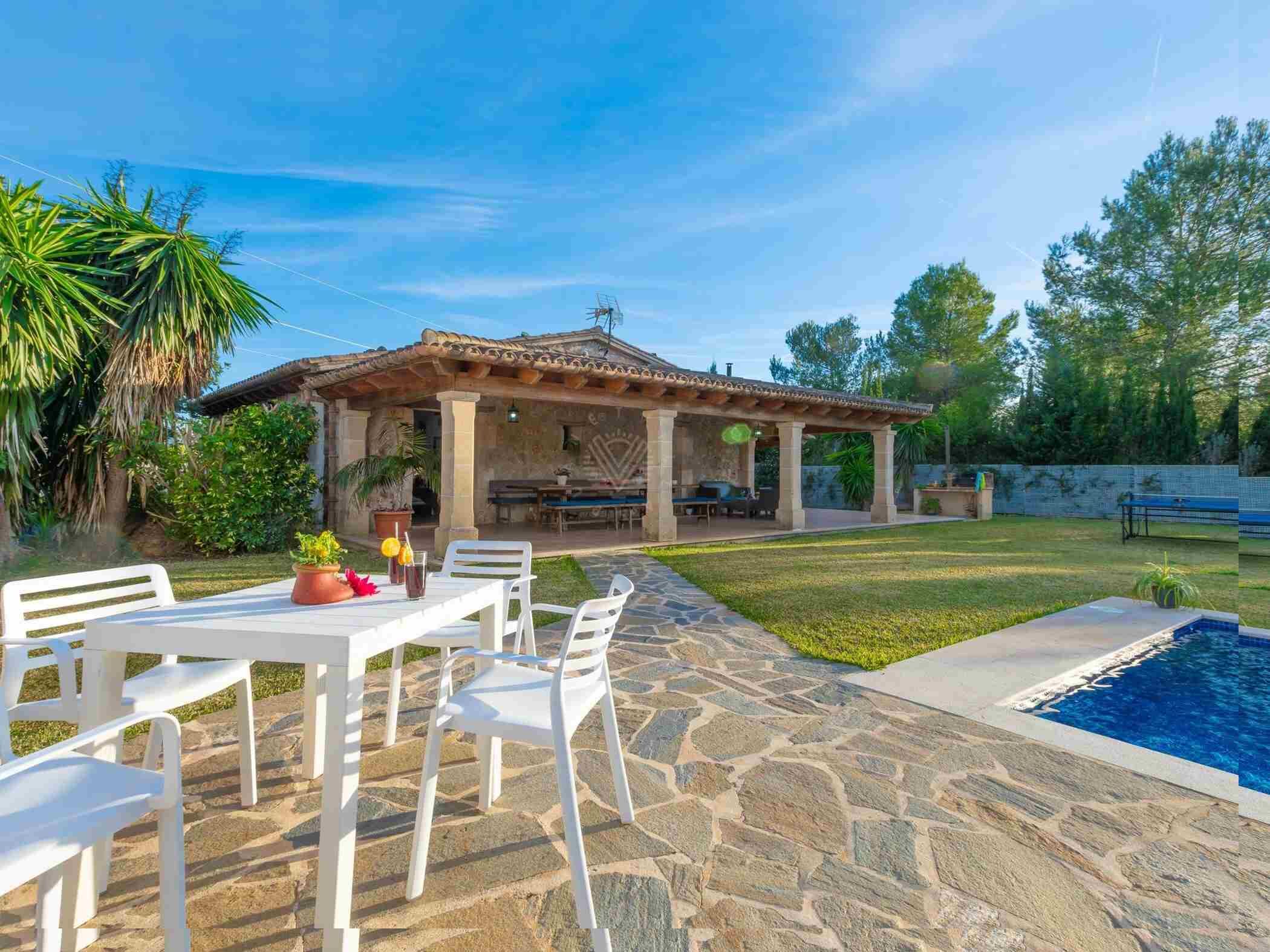 Un patio con suelo de piedra, sillas blancas, una mesa y una piscina.