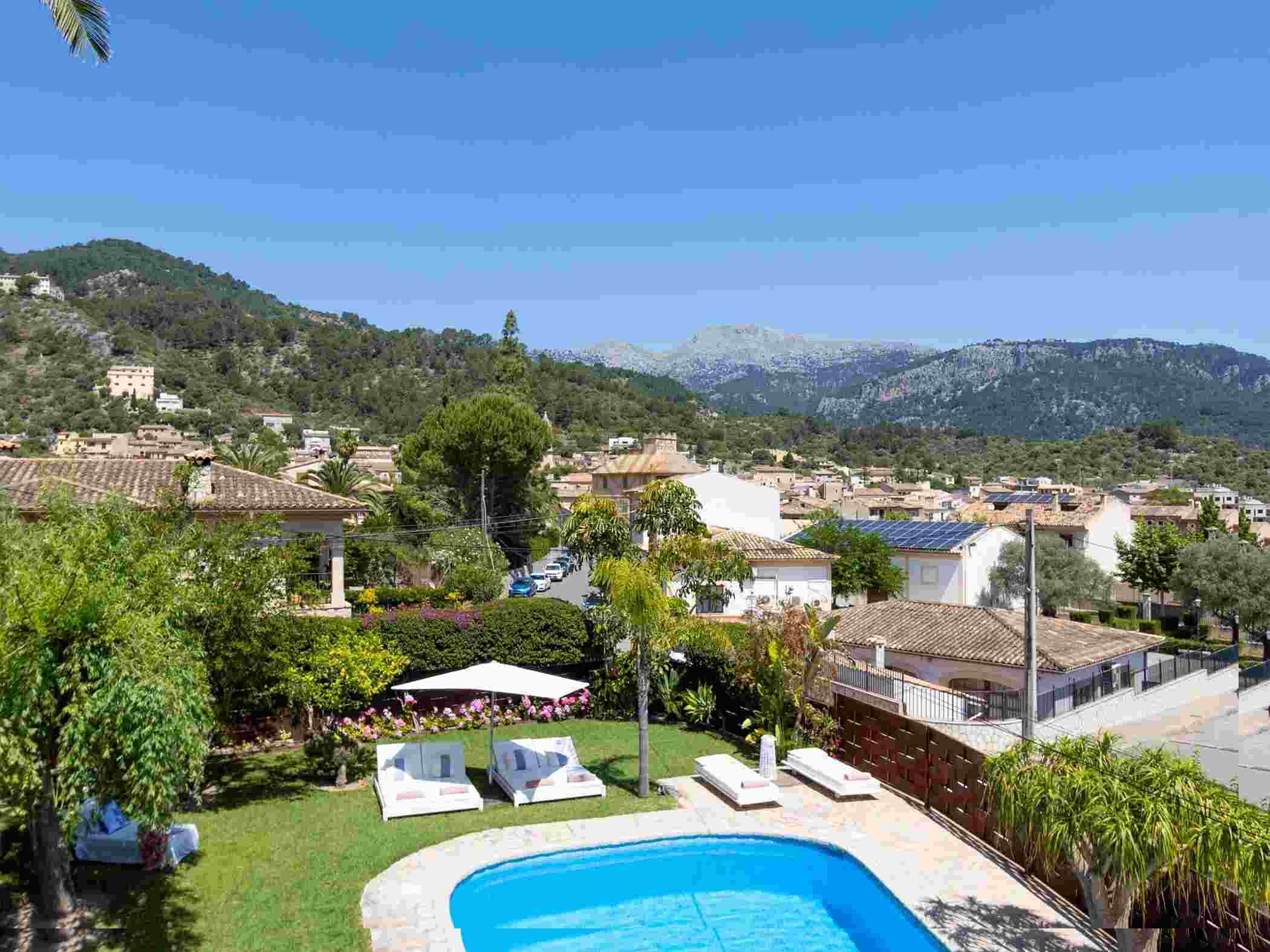 A pool area with lounge chairs, a garden, and a view of a town with buildings and mountains in the background.