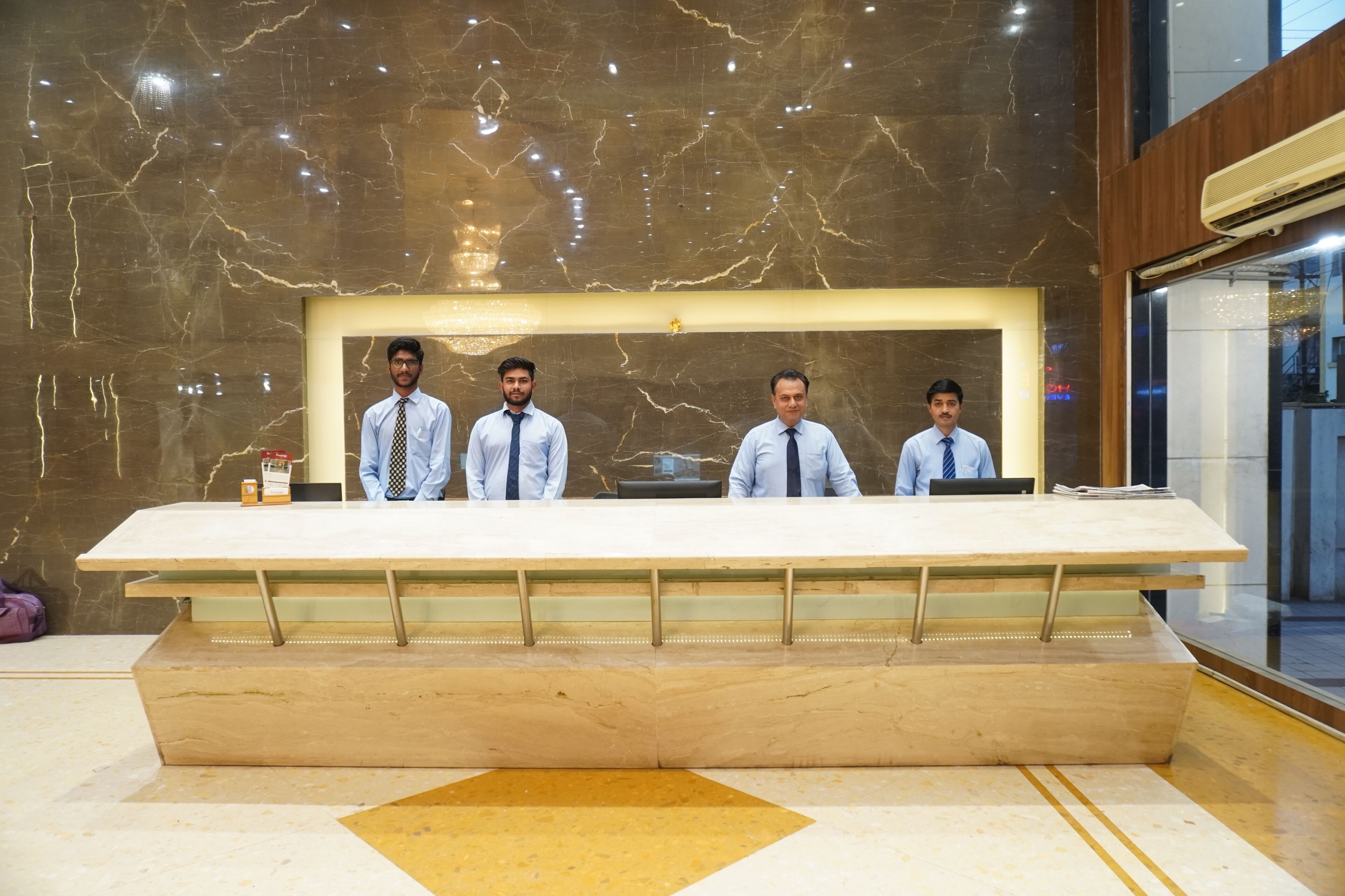 A reception desk with four individuals seated behind it, set against a marble wall with a geometric pattern.