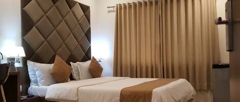 Une chambre d’hôtel avec un grand lit, une table de chevet en verre, une tête de lit moderne, des rideaux beiges et un appareil de climatisation.