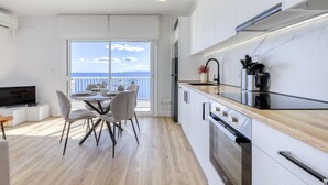 A modern kitchen with a large window overlooking the sea, a dining area with a table and chairs, and a TV on a white cabinet.