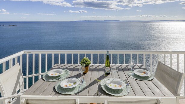 A table set for four with plates, cutlery, and a bottle of wine, overlooking a calm sea with distant land.