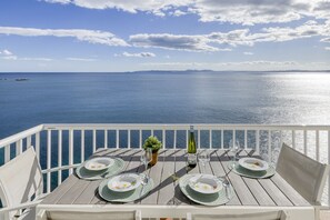 A table set for four with plates, cutlery, and a bottle of wine, overlooking a calm sea with distant land.