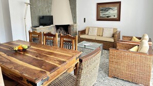 A dining area with a wooden table, chairs, and a fruit basket. There is a fireplace, a sofa, wicker furniture, and an air conditioning unit on the wall. - Agi Joan Badosa 2 Apartamento for rent in Roses Girona. (Roses)