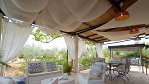 An outdoor patio with wicker furniture, a stone floor, and a view of olive trees. - Figuera Blanca (Buger)