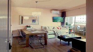 A modern living room with a sofa, a dining table with chairs, a TV mounted on the wall, and a large window providing natural light. - QUAD 2 with Balcony, Garage, A.C. (Cold or heat), 24/7 security  (Lisboa)