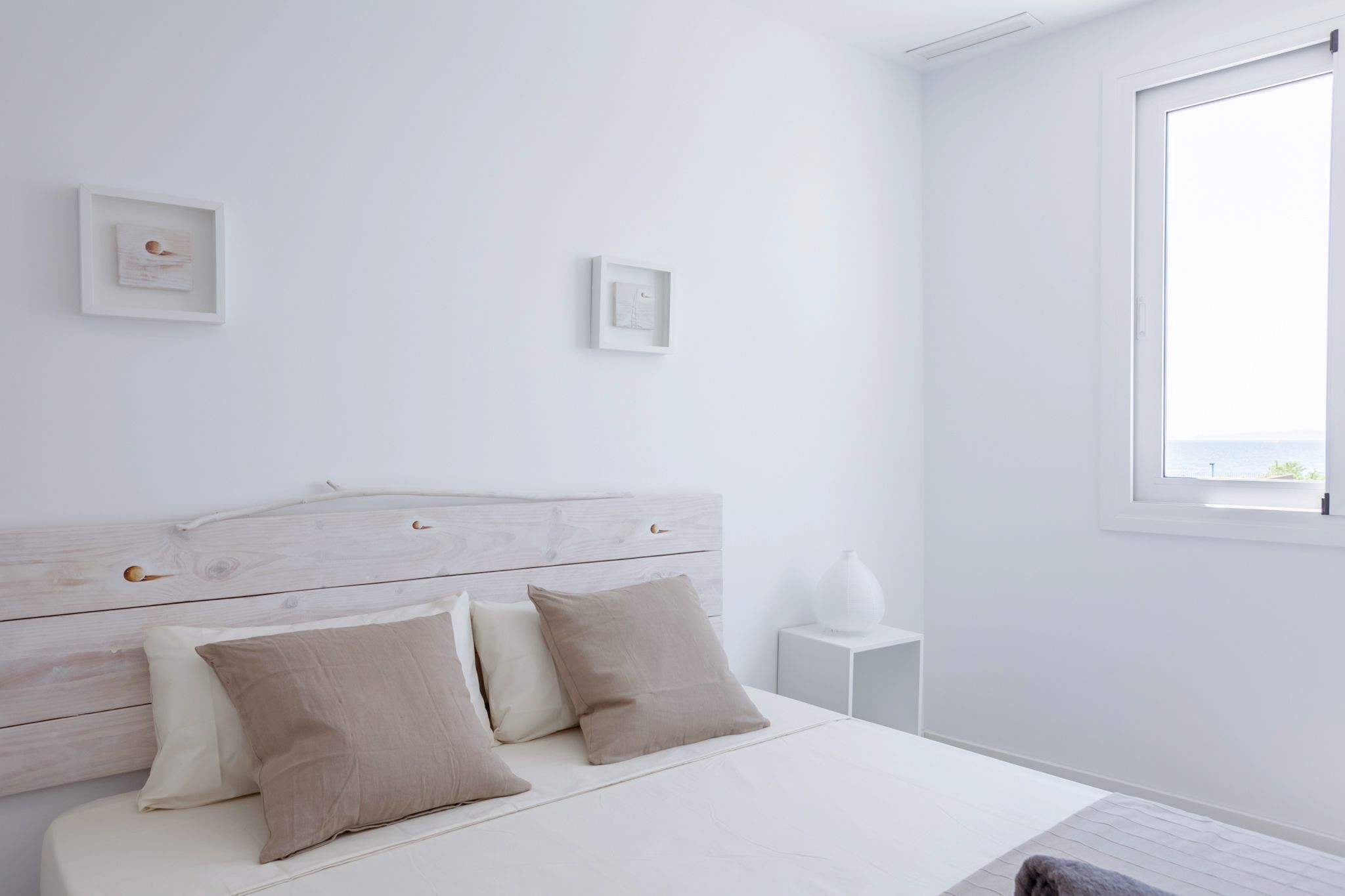 A minimalist bedroom with a white bed, a wooden headboard, two bedside tables, and a window with a view.