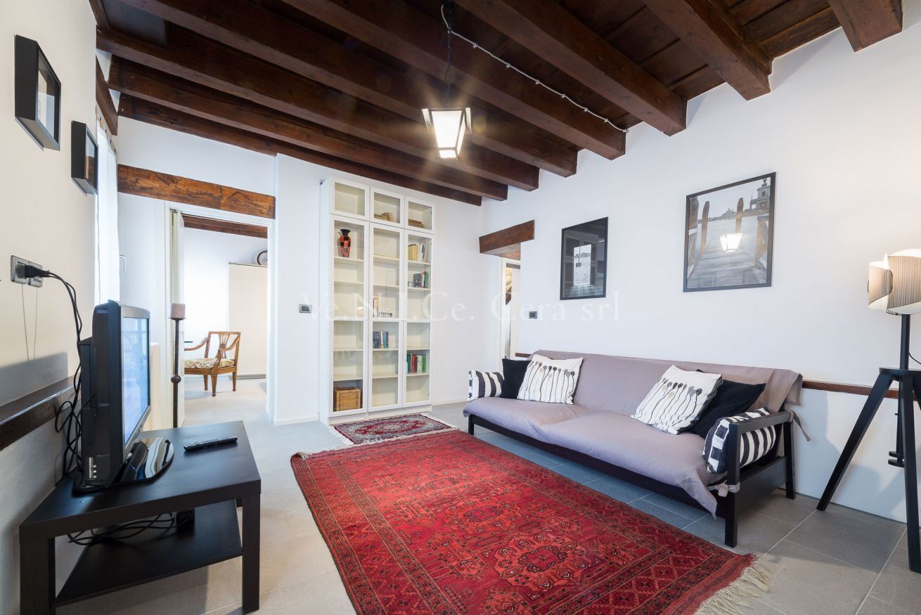Una sala de estar con un sofá, una alfombra roja, un televisor sobre un mueble bajo y una estantería.
