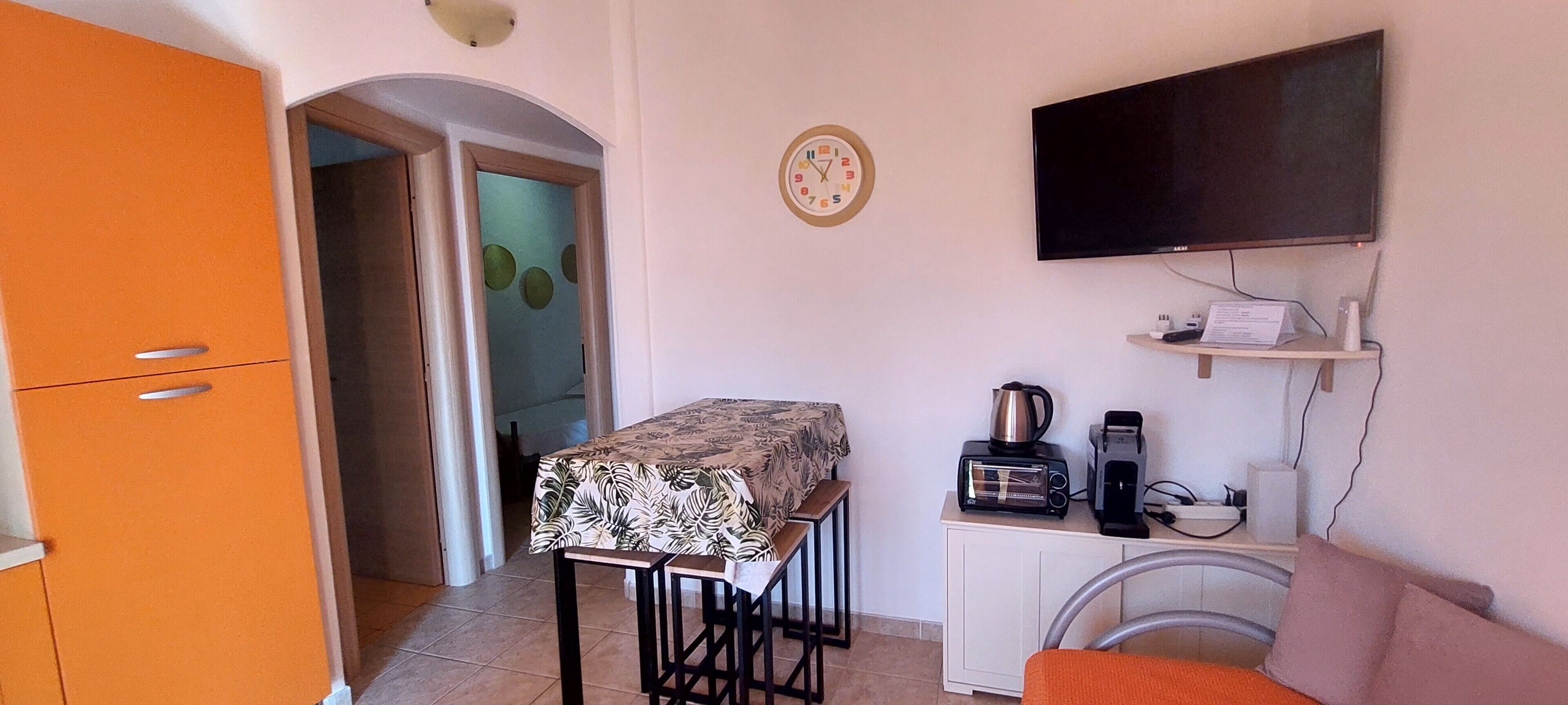 Une pièce compacte comprenant un coin cuisine, un téléviseur fixé au mur, une horloge et une petite table recouverte d’une nappe.