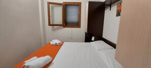 Een klein, net opgemaakt bed met witte lakens en een oranje dekbed, een houten hoofdbord, een klein raam met zonwering en een spiegel aan de muur.