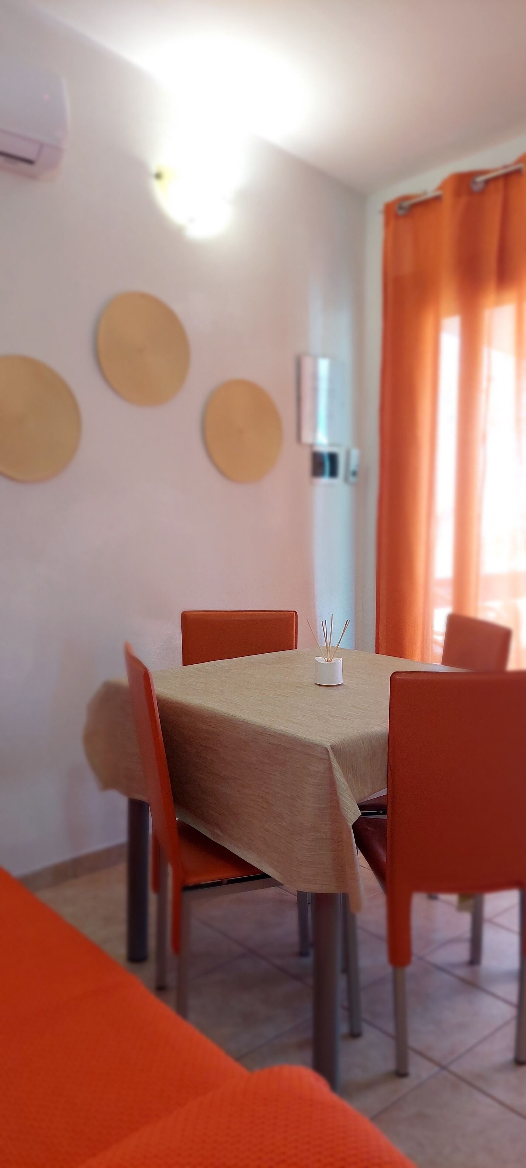 Een eethoek met een tafel bekleed met een beige tafelkleed, omringd door oranje stoelen. Drie ronde decoraties hangen aan de muur, evenals een airconditioner.
