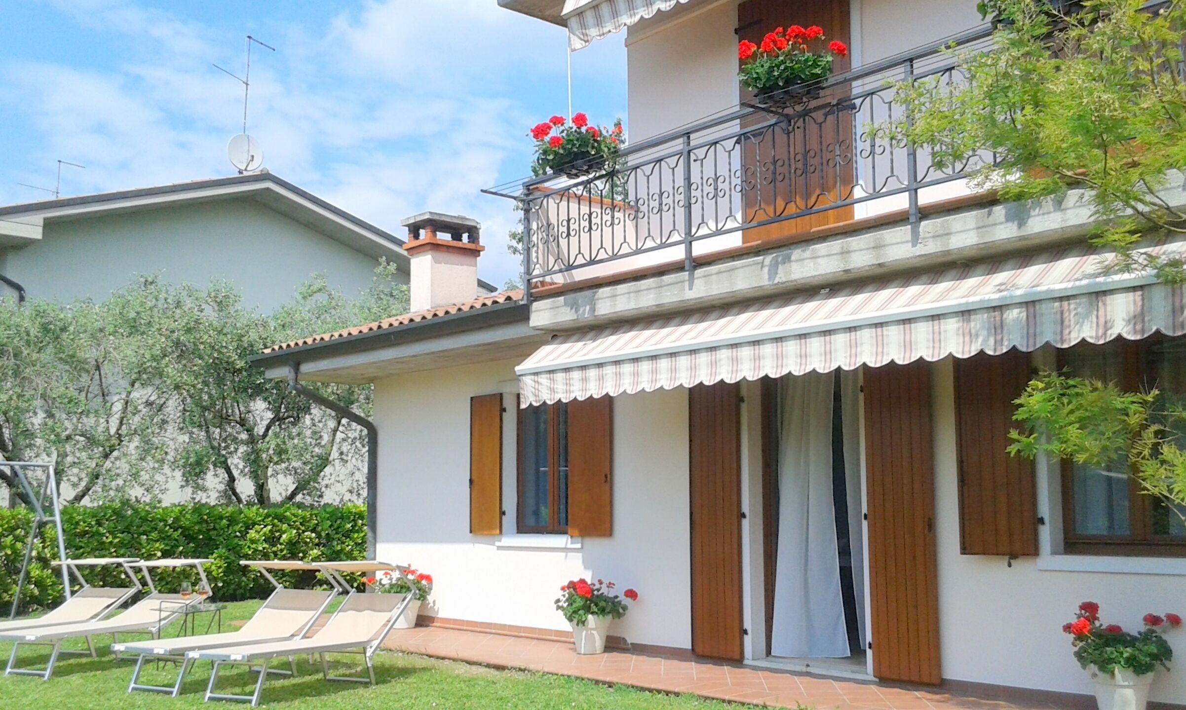 A two-story house with a balcony, a covered porch, and a garden with sun loungers.
