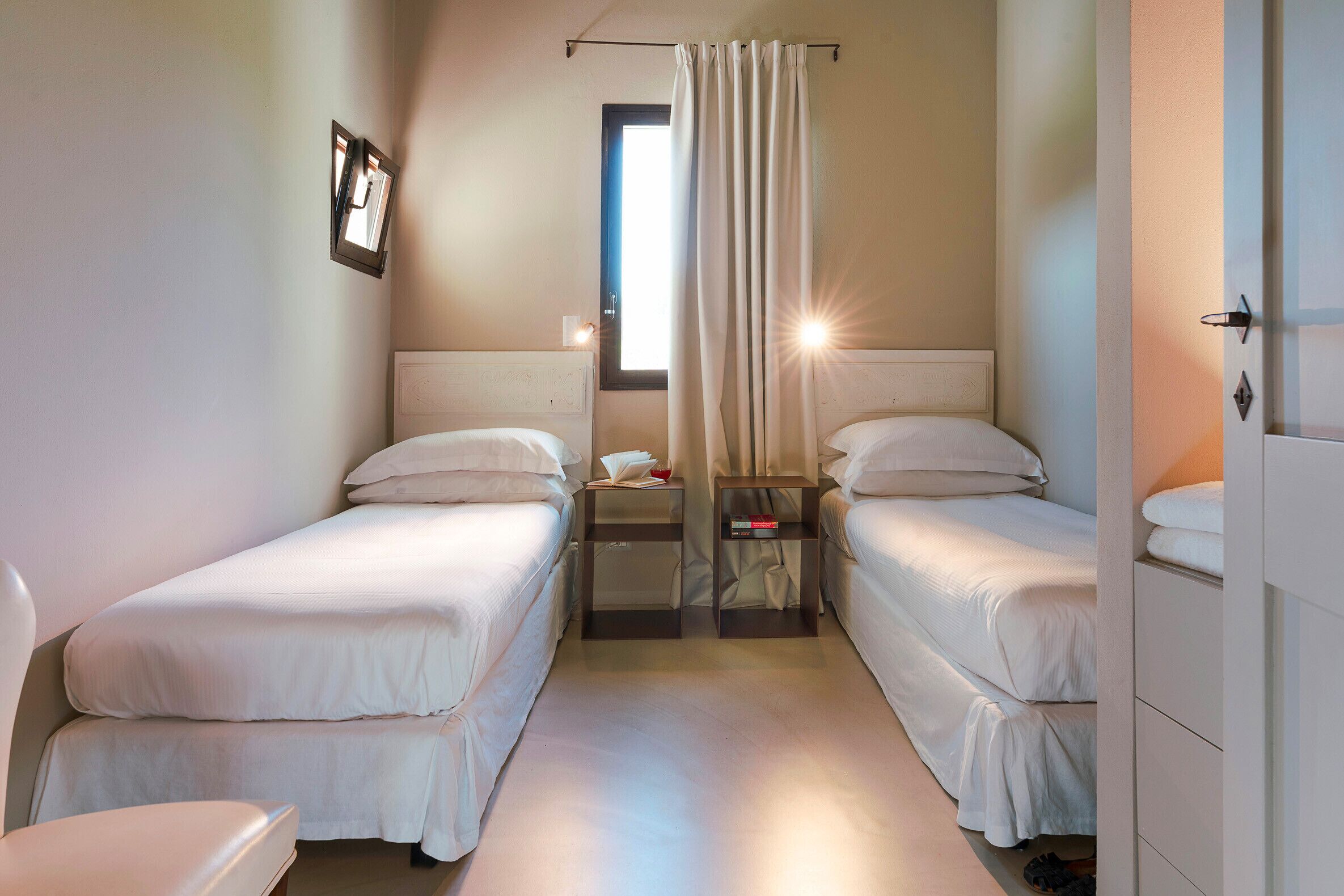 Une chambre d’hôtel avec deux lits simples, une petite table avec une lampe, une fenêtre avec des rideaux et une porte avec une poignée.