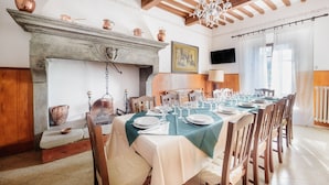 Zona de dinar amb una taula llarga preparada per a un àpat, una llar de foc amb olins de coure, bigues de fusta i un penjador.