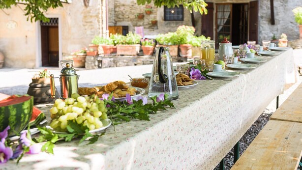 Una taula llarga a l'aire lliure, plena d'aliments i begudes, envoltada de plantes en testos i un edifici de pedra al fons.