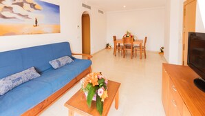 A living room with a blue sofa, a wooden coffee table with a floral arrangement, a dining area with a wooden table and chairs, and a TV on a wooden cabinet. - Nice apartment with terrace and pool near the beach and shops Inmosantos Daniel AticB (Rosas / Roses)