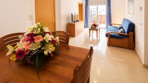 A dining area with a wooden table and chairs, a floral centerpiece, a sofa, and a television on a wooden cabinet. - Nice apartment with terrace and pool near the beach and shops Inmosantos Daniel AticB (Rosas / Roses)