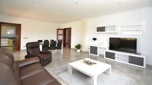 Una sala moderna con un televisor de pantalla plana grande, sofás de cuero, una zona de comedor con sillas y una cocina al fondo.
