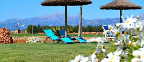 A grassy area with two thatched umbrellas, a windmill, and a mountain backdrop.