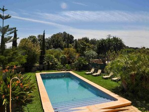 Une piscine rectangulaire bordée d’une terrasse en bois, entourée d’une végétation luxuriante et sous un ciel dégagé.