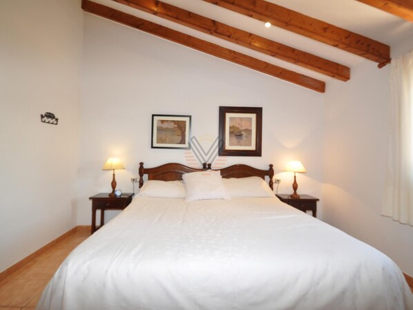 Une chambre avec un grand lit, des poutres en bois au plafond, deux tables de chevet avec des lampes et deux tableaux encadrés au mur.