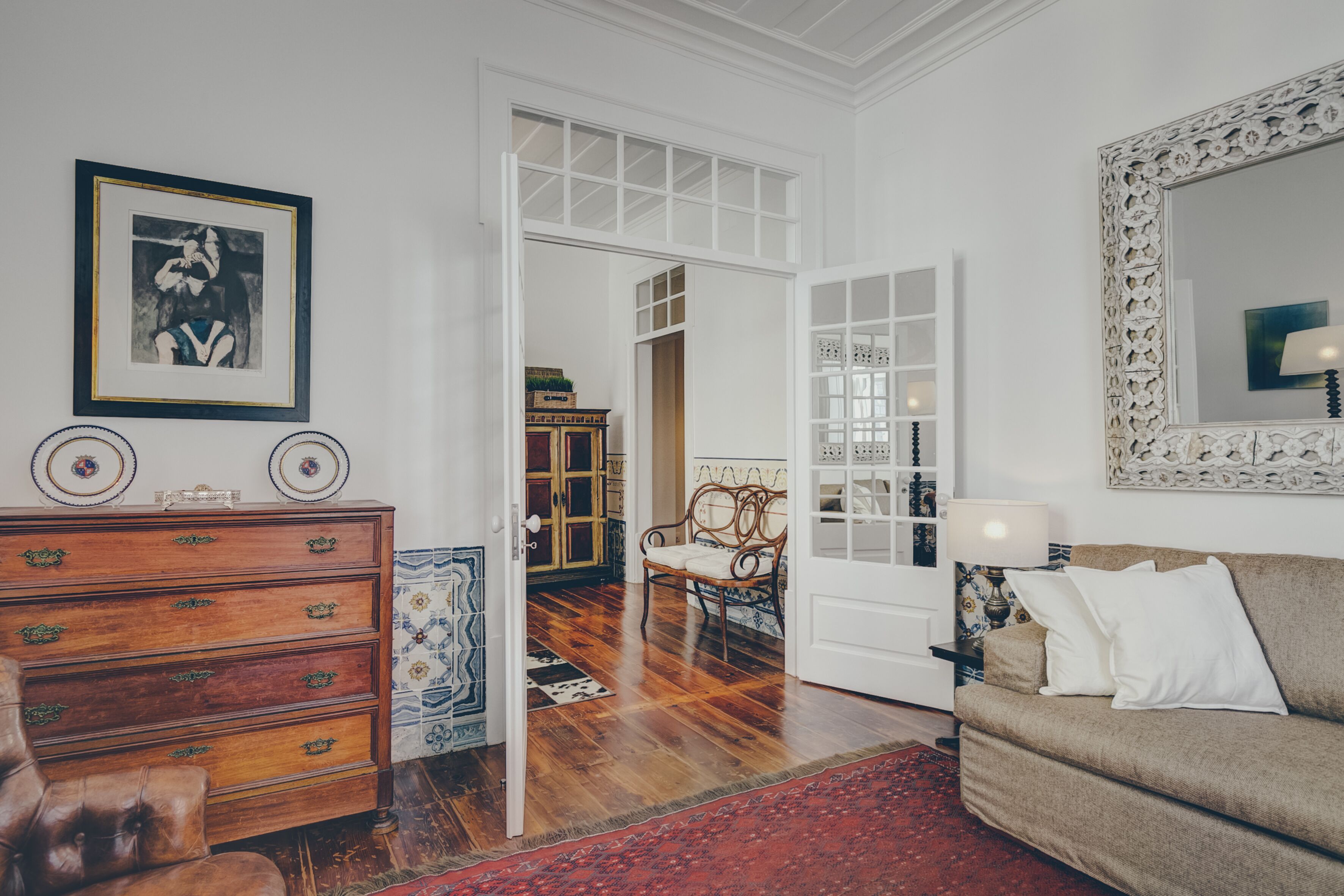 Una habitación con un aparador de madera, un sofá, una silla y un espejo grande. Hay una puerta que da a otra habitación.