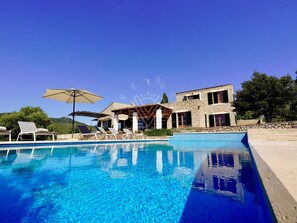 Uma piscina com espreguiçadeiras e guarda-sóis, um edifício de pedra com várias janelas, e um céu azul límpido.