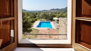 A wooden-framed window with a view of a swimming pool, stone walls, and a distant landscape. - Charming Mallorcan house with beautiful views (Son Servera)