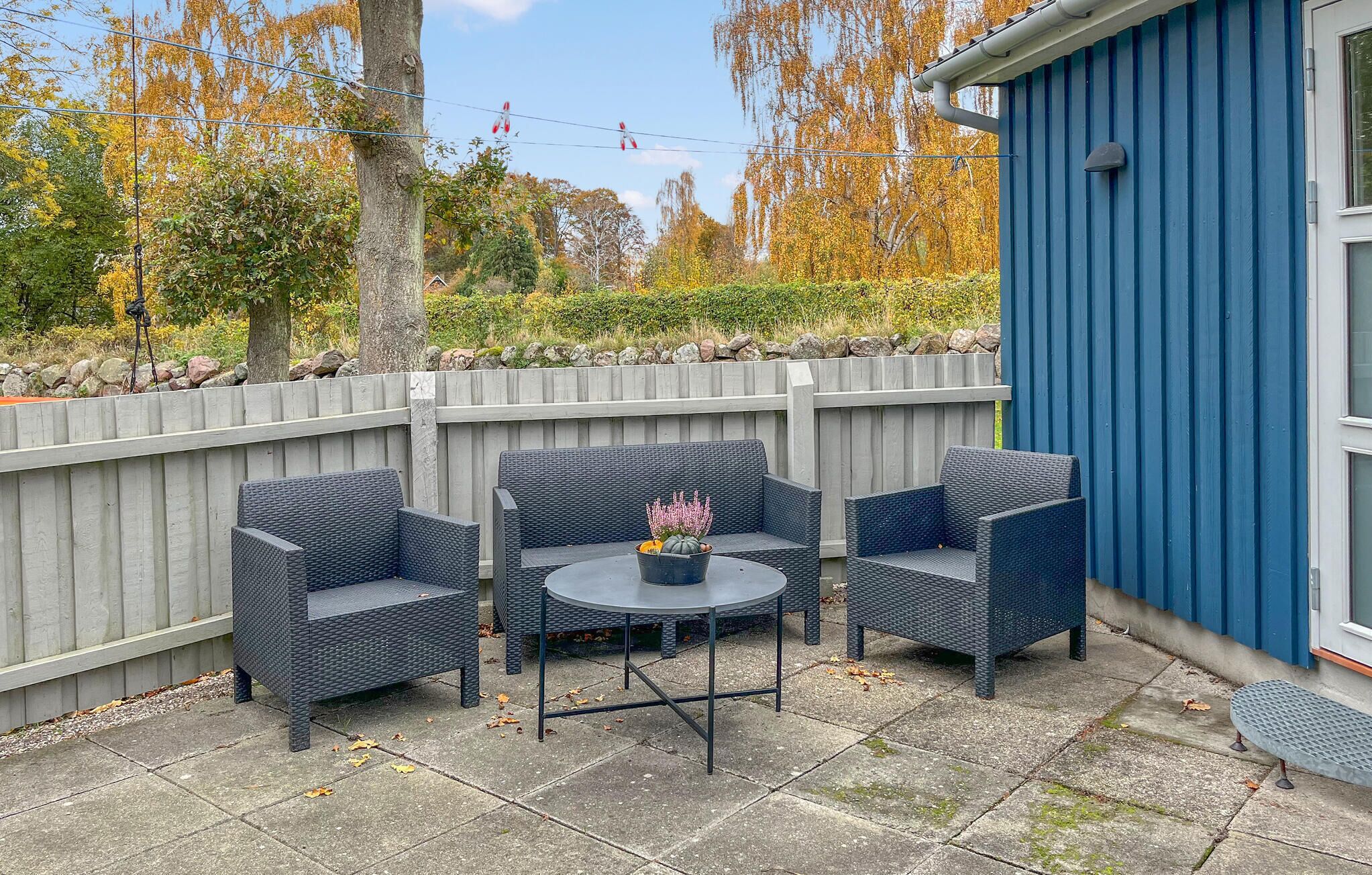 Eine Terrasse im Freien mit Korbmöbeln und einem kleinen Tisch, umgeben von einem Holzzaun und einem blauen Gebäude.