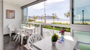 A modern dining area with white chairs and tables, large windows with a view of the beach and ocean, and a potted plant on the table. - RESIDENCE DE LA PLAGE BJS 3 Roses (Rosas / Roses)