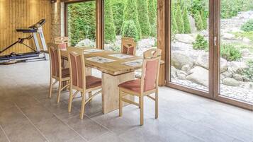 Ein Essbereich mit Holztisch und -stühlen, großen Fenstern und Trainingsgeräten im Hintergrund.
