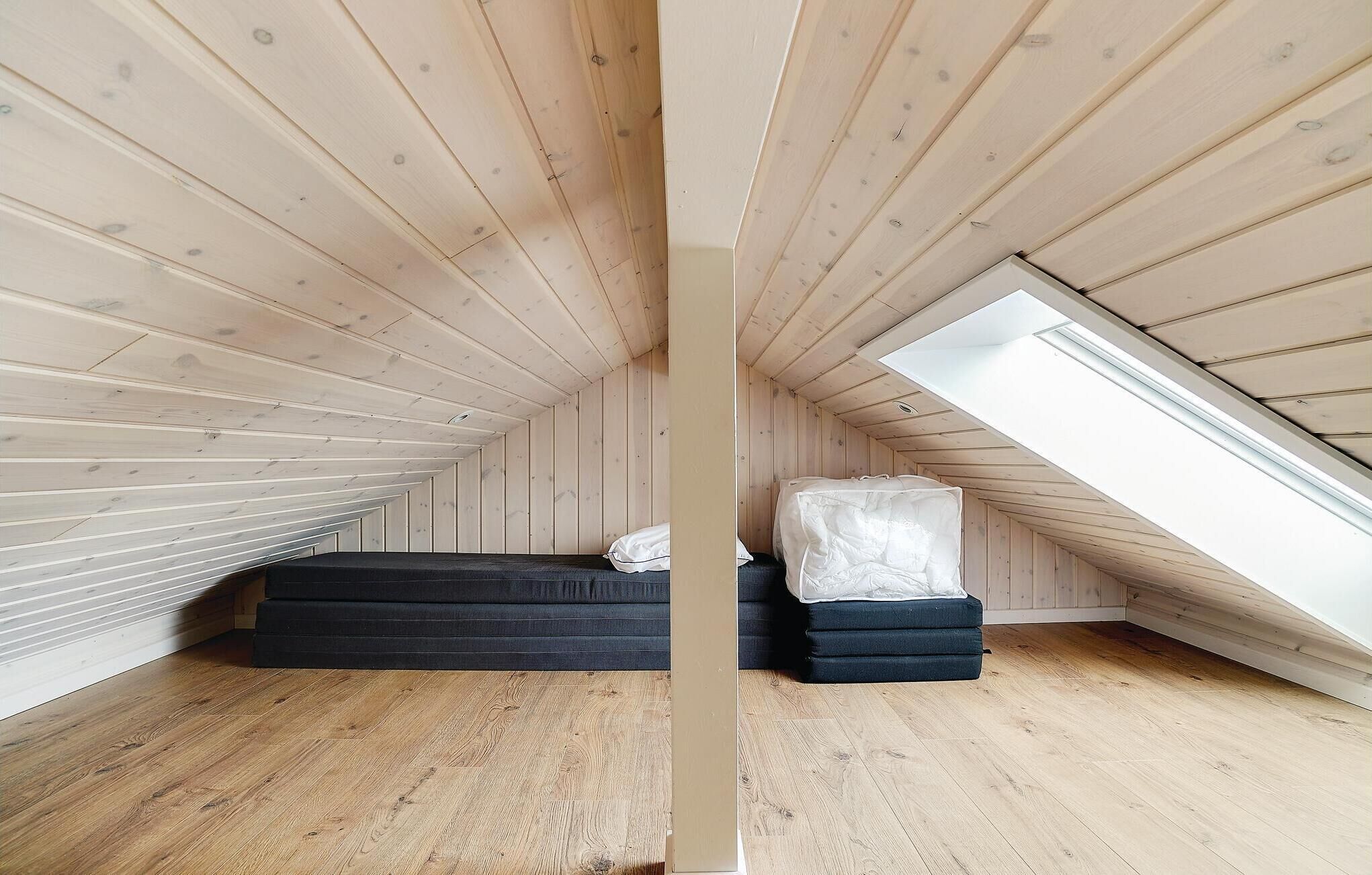 Ein kleines Schlafzimmer im Dachgeschoss mit Holzwänden und einer geneigten Decke. Es gibt zwei Betten mit Matratzen und Kissen sowie ein Oberlicht auf der rechten Seite.