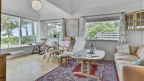 Et lyst rum med trægulv, et rundt bord, en sofa og to stole. Der er vinduer med gardiner, en lampe og et kort på væggen.