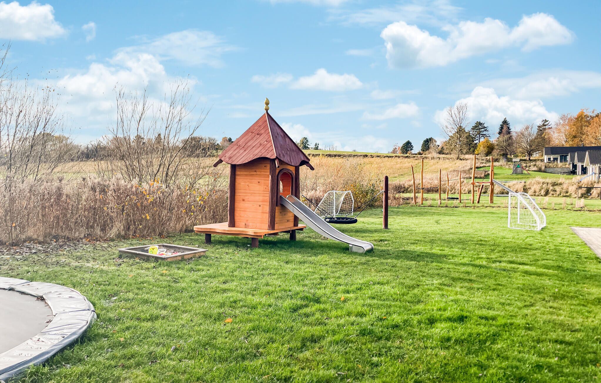 A wooden playhouse with a slide, surrounded by a grassy field and a pond.
