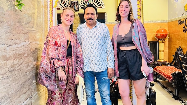 A group of people standing in a room with ornate floral and geometric wall designs, a patterned floor, and a bed with a red and black patterned bedspread. - Hotel Moon Light Palace Jaipur (Jaipur)