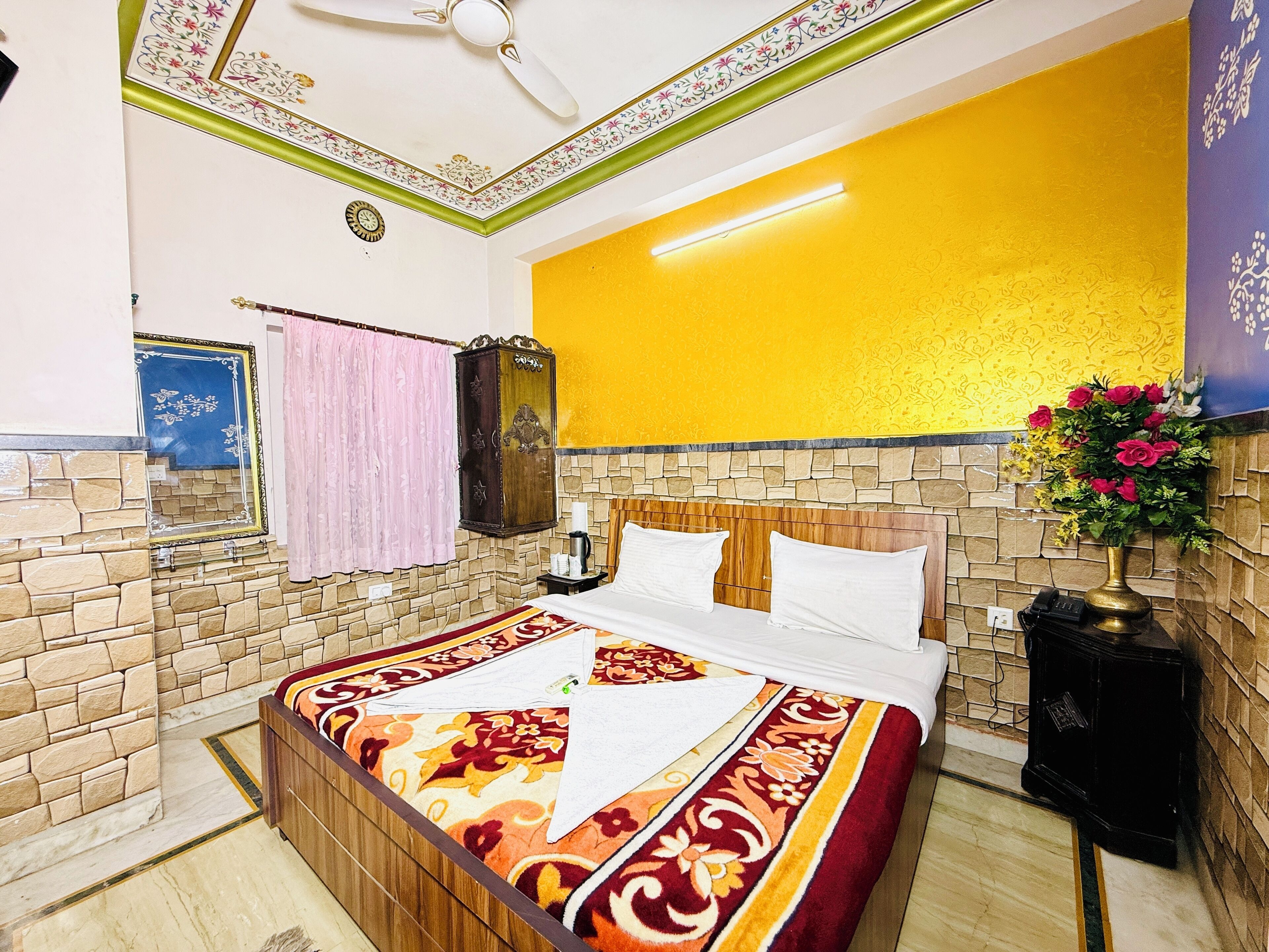 A hotel room with a bed, a nightstand, a mirror, a clock, a ceiling fan, and a vase with flowers.